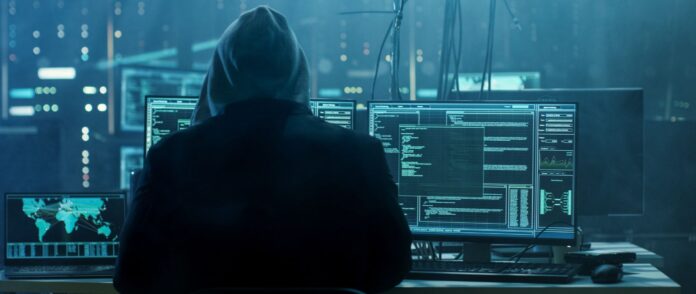 Sicherheitsrisiko: Ein Hacker mit Kapuze sitzt vor mehreren Bildschirmen.