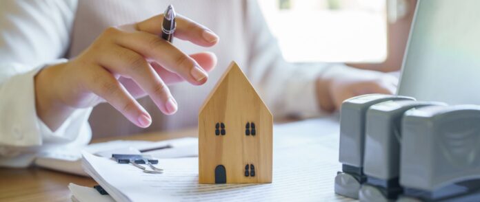 Immobilienverwaltung: Eine Frau arbeitet im Hintergrund und hält einen Stift über einem Holzhaus, welches auf einem Stapel Papier steht.