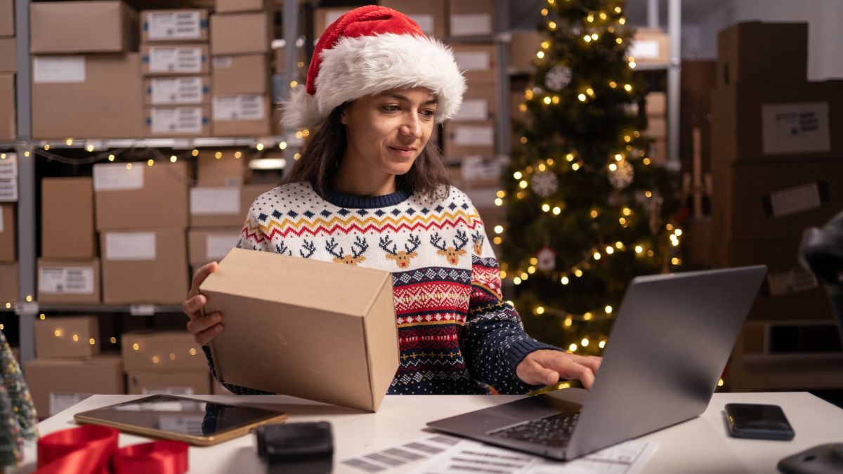 Weihnachtsgeschäft: Kleinunternehmerin, die am Laptop arbeitet und Pakete für die Auslieferung am Heiligabend vorbereitet und verpackt.