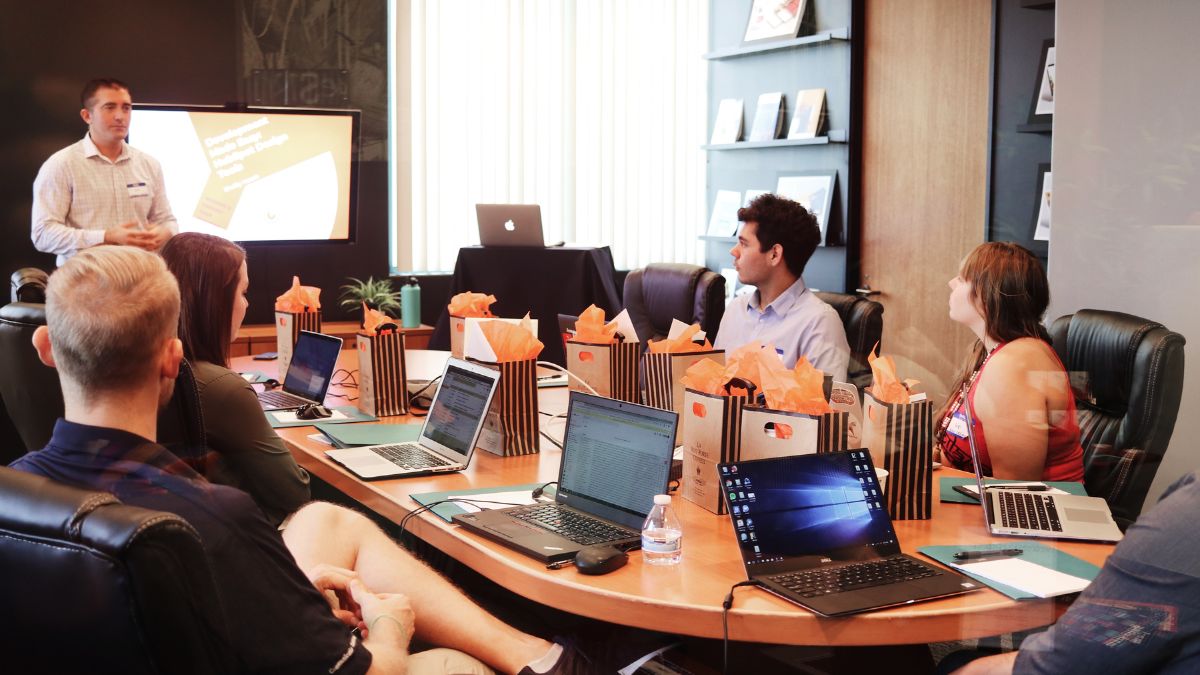 Hybride Meetings: Ein Mann steht vor Menschen, die an einem Tisch mit Laptops sitzen.