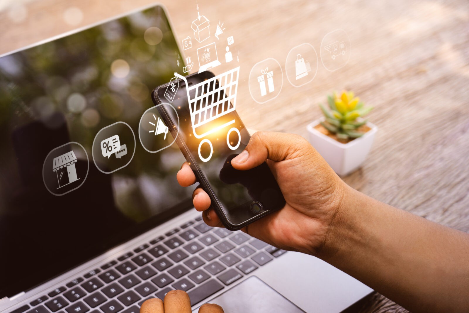Symbolbild E-Commerce: Laptop und Smartphone in einer Hand mit Einkaufssymbolik